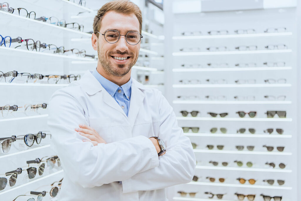Óptico sonriente con gafas y bata blanca en tienda de monturas, atención profesional en Óptica Giner.